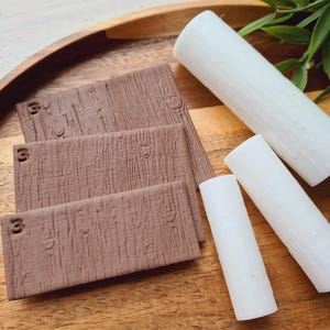Könnte beinhalten: Auf einem Holztablett liegen drei rechteckige, braune Blöcke mit Holzmaserung und der eingravierten Zahl "3". Daneben befinden sich drei weiße zylindrische Objekte. Grüne Blätter sind im Hintergrund.