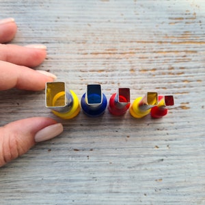 May include: A set of five metal and plastic square-shaped cutters in varying sizes. The cutters have a silver metal cutting edge and are mounted on colorful plastic bases of yellow, blue, and red. The cutters are arranged in a row.
