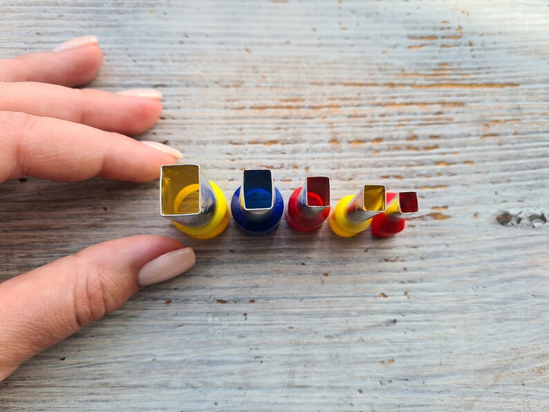 May include: A set of five metal and plastic square-shaped cutters in varying sizes. The cutters have a silver metal cutting edge and are mounted on colorful plastic bases of yellow, blue, and red. The cutters are arranged in a row.