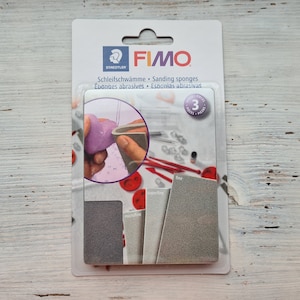 May include: A pack of FIMO sanding sponges for polymer clay. The pack shows three sponges: microfine, superfine, and fine. The pack also includes a picture of a hand sanding a purple heart-shaped piece of clay.