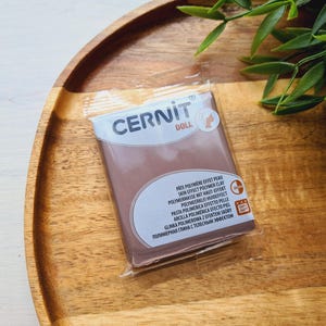 May include: A package of Cernit Doll polymer clay in a skin effect color. The package is sealed in clear plastic and sits on a wooden tray. The text on the package includes "Skin Effect Polymer Clay".