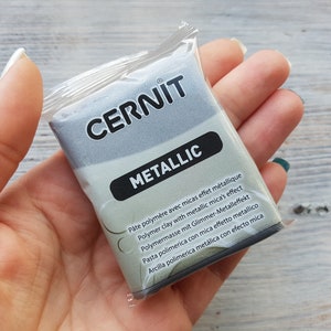 Puede incluir: Un paquete de arcilla polim&eacute;rica gris metalizado con el nombre de la marca "CERNIT" y el texto "METALLIC" impreso en la parte delantera. El paquete incluye instrucciones en varios idiomas.