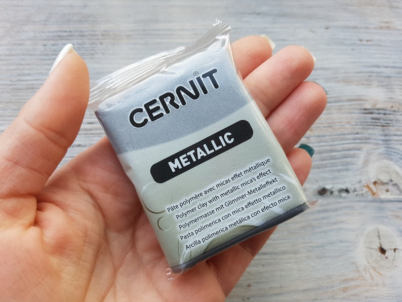 Puede incluir: Un paquete de arcilla polim&eacute;rica gris metalizado con el nombre de la marca "CERNIT" y el texto "METALLIC" impreso en la parte delantera. El paquete incluye instrucciones en varios idiomas.