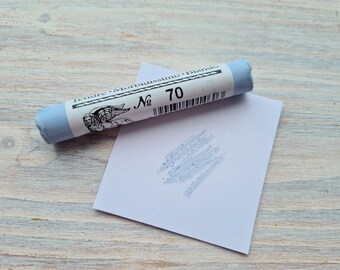 May include: A light blue pastel stick labeled "No 70" with a sample stroke on a white paper. The pastel is cylindrical with a white label featuring text and an illustration of a winged figure. The background is a light wood surface.