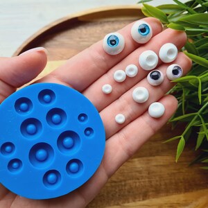 K&ouml;nnte beinhalten: Eine blaue Silikonform mit 10 runden Vertiefungen zum Herstellen von Puppenaugen. Die Form liegt auf einer Hand, und auf der Hand befinden sich mehrere Puppenaugen in verschiedenen Gr&ouml;&szlig;en und Farben.