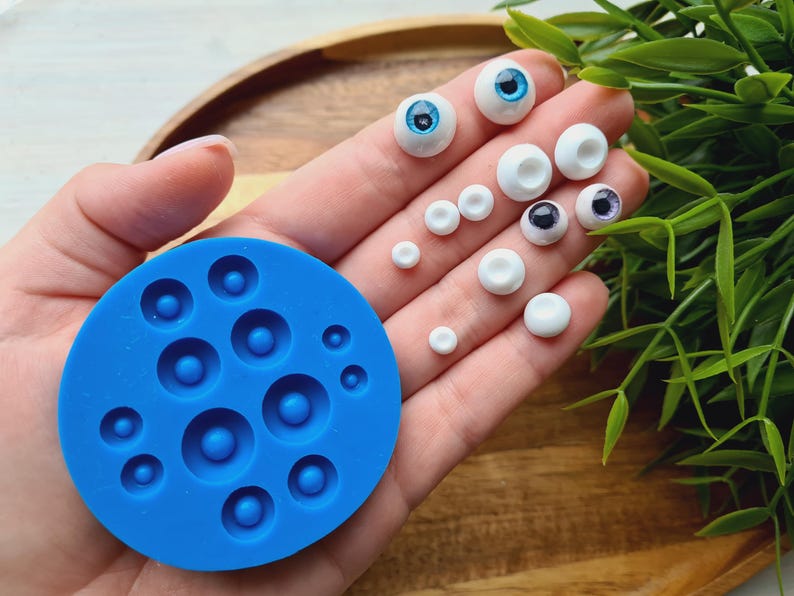 K&ouml;nnte beinhalten: Eine blaue Silikonform mit 10 runden Vertiefungen zum Herstellen von Puppenaugen. Die Form liegt auf einer Hand, und auf der Hand befinden sich mehrere Puppenaugen in verschiedenen Gr&ouml;&szlig;en und Farben.