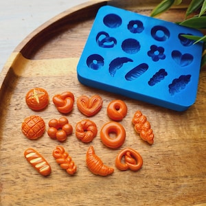 Op de afbeelding: Oranjegekleurde miniatuurvormen voor brood en gebak en afgewerkte producten. Een blauwe siliconen mal met verschillende vormen staat naast de afgewerkte producten. De items zijn gerangschikt op een houten dienblad. De afgewerkte producten zijn onder meer een pretzel, croissant en donut.