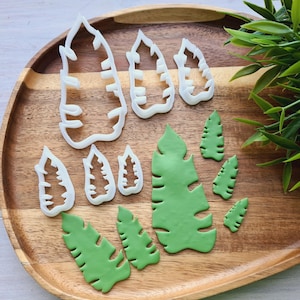 May include: A set of white plastic leaf-shaped cookie cutters and green fondant leaves on a wooden surface.