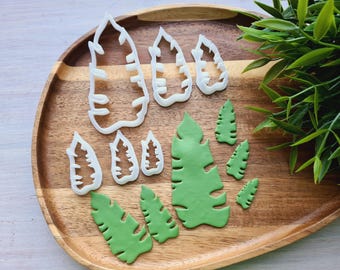 May include: A set of white plastic leaf-shaped cookie cutters and green fondant leaves on a wooden surface.