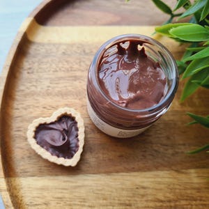 Könnte beinhalten: Ein Glas gefüllt mit dunkelbrauner Schokoladencreme auf einem Holztablett mit einem herzförmigen Keks, gefüllt mit Schokoladencreme.