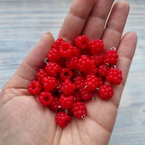 Puede incluir: Una mano sosteniendo un montón de abalorios en forma de frambuesa roja con anillos de salto plateados.