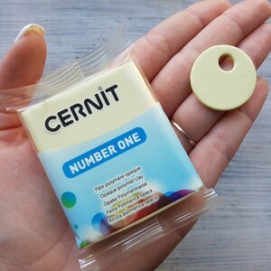 May include: A yellow package of CERNIT NUMBER ONE opaque polymer clay. The package is in a person's hand, and a small yellow disc of clay is next to it.
