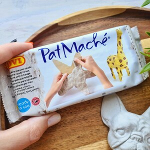 Pode incluir: Uma embalagem branca de massa de papel mach&eacute; Pat Mach&eacute; com uma girafa e uma escultura em forma de m&atilde;o na frente. A embalagem diz "pronto a usar" e tem um logotipo azul e branco.
