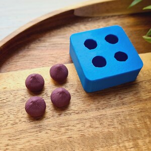 Op de afbeelding: Vier kleine, donkerpaarse, koepelvormige snoepjes liggen op een houten oppervlak naast een blauwe siliconen mal met vier ronde holtes.