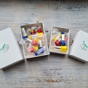 May include: Four white boxes with the text "Polymer Clay" on a sticker. Two boxes are open, revealing colorful polymer clay tools in red, yellow, and blue. The tools are packaged in clear plastic. The boxes are on a weathered, light-colored wooden surface.