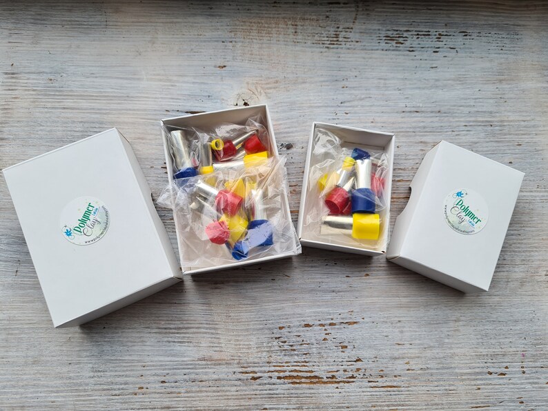 May include: Four white boxes with the text "Polymer Clay" on a sticker. Two boxes are open, revealing colorful polymer clay tools in red, yellow, and blue. The tools are packaged in clear plastic. The boxes are on a weathered, light-colored wooden surface.