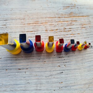 May include: A set of eight metal and plastic punch tools of varying sizes, arranged in a row. Each tool has a square opening and a colored plastic base, including yellow, blue, and red. The tools are on a light wood surface.