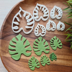 May include: A set of eight white plastic cookie cutters shaped like monstera leaves, arranged on a wooden surface with green fondant monstera leaves.