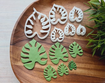 May include: A set of eight white plastic cookie cutters shaped like monstera leaves, arranged on a wooden surface with green fondant monstera leaves.