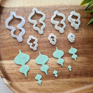 Puede incluir: Un juego de cortadores de galletas de pl&aacute;stico blanco con forma de hojas y un juego de galletas verdes con forma de hojas sobre una superficie de madera.