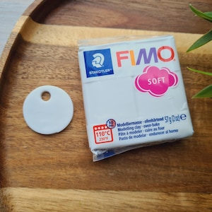 May include: A white FIMO Soft modeling clay package with a pink cloud-shaped "SOFT" label, a white ceramic tag with a hole, and green leaves on a wooden tray. The package indicates oven-bake at 230&deg;F (110&deg;C).