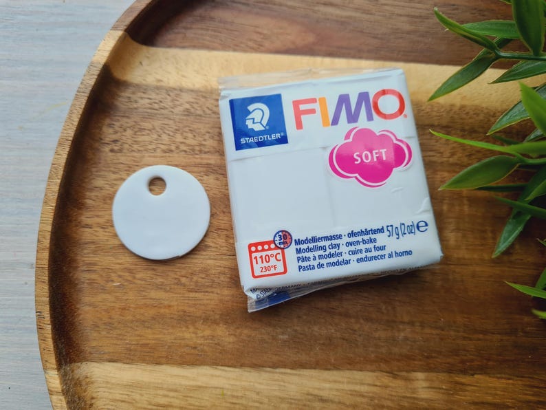 May include: A white FIMO Soft modeling clay package with a pink cloud-shaped "SOFT" label, a white ceramic tag with a hole, and green leaves on a wooden tray. The package indicates oven-bake at 230&deg;F (110&deg;C).