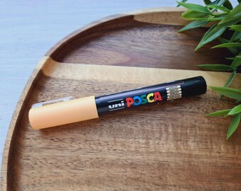 May include: A yellow Uni Posca paint marker on a wooden surface. The marker has black lettering and a black cap.