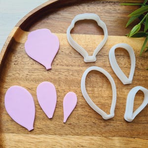 Op de afbeelding: Een verzameling bloemvormige koekjessnijders en uitgesneden fondantstukken in verschillende maten en vormen. De uitstekers zijn wit en de fondantstukken lichtpaars. De items zijn gerangschikt op een houten dienblad.