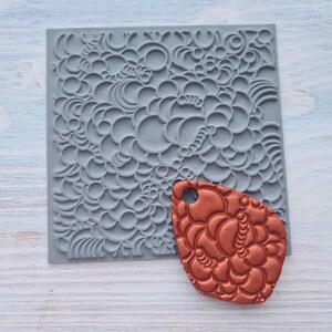 Puede incluir: Una estera de estampado en silicona gris con un patrón de círculos superpuestos y una pieza de arcilla estampada de color cobre.