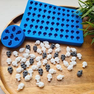 Puede incluir: Una bandeja de madera contiene un molde de silicona azul con cavidades en forma de calavera, junto con objetos dispersos en forma de calavera blancos y grises. También hay un molde circular con formas de calavera.