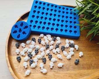May include: A rectangular blue silicone mold with many small skull cavities and a circular blue silicone mold with seven skull cavities, accompanied by a collection of small, individual white and dark gray skull-shaped pieces.