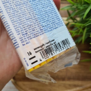 Puede incluir: Primer plano de un paquete de arcilla polim&eacute;rica FIMO en un envoltorio de pl&aacute;stico transparente. La etiqueta muestra el nombre del color "amarillo sol" y un c&oacute;digo de barras. El fondo incluye follaje verde.