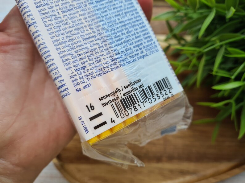 Puede incluir: Primer plano de un paquete de arcilla polim&eacute;rica FIMO en un envoltorio de pl&aacute;stico transparente. La etiqueta muestra el nombre del color "amarillo sol" y un c&oacute;digo de barras. El fondo incluye follaje verde.