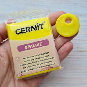 Może przedstawiać: Żółte opakowanie gliny polimerowej CERNIT OPALINE z ceramicznym wykończeniem. Opakowanie jest oznaczone w wielu językach. Obok opakowania znajduje się mały żółty dysk z gliny z otworem w środku.