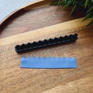 May include: A black, zig-zag shaped tool and a light blue, zig-zag shaped piece sit on a wooden surface. The tool is made of a hard material, and the blue piece appears to be a soft material. The items are likely used for crafting or baking.