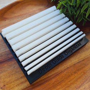 Peut inclure: Un organisateur de guide de profondeur noir contenant plusieurs guides de profondeur blancs. L'organisateur est rectangulaire et repose sur un plateau en bois. Le texte "Depth Guide Organizer" est imprim&eacute; sur l'organisateur noir.