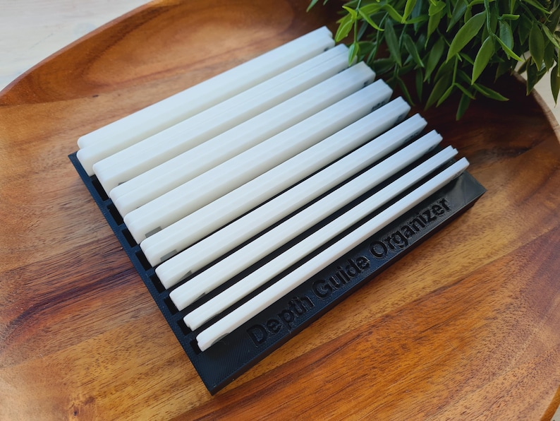 Peut inclure: Un organisateur de guide de profondeur noir contenant plusieurs guides de profondeur blancs. L'organisateur est rectangulaire et repose sur un plateau en bois. Le texte "Depth Guide Organizer" est imprim&eacute; sur l'organisateur noir.