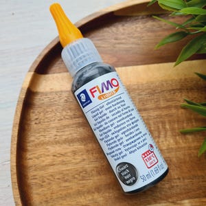 Op de afbeelding: Een zwarte fles FIMO Liquid, een heldere ovenhardende gel voor polymer clay. De fles is voorzien van instructies in meerdere talen en heeft een zwarte dop met een gele bovenkant.