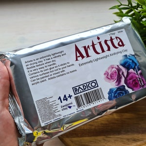 May include: A silver package of white air-drying clay labeled "Artista" with a barcode and the text "Extremely Lightweight Airdrying Clay". The package features a floral design with pink, purple, and blue roses. The package also includes the text "170gr White" and "14+ Zwajomi clay".