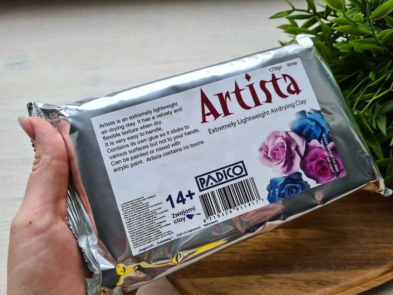 May include: A silver package of white air-drying clay labeled "Artista" with a barcode and the text "Extremely Lightweight Airdrying Clay". The package features a floral design with pink, purple, and blue roses. The package also includes the text "170gr White" and "14+ Zwajomi clay".