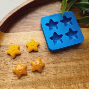 Pode incluir: Uma bandeja de madeira contém um molde de silicone azul com quatro cavidades em forma de estrela e vários doces amarelos em forma de estrela. Os doces têm rostos sorridentes. A bandeja está sobre uma superfície de madeira.