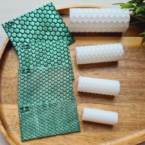 May include: A wooden tray displays various items. On the left are green, scale-patterned fabric pieces. To the right are four white, honeycomb-textured wax rolls. The tray is brown, and a small green plant is in the background.
