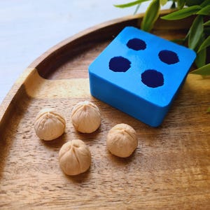 Pode incluir: Quatro pequenos objetos redondos, lisos e de cor marrom claro sobre uma superfície de madeira ao lado de uma forma de silicone azul com quatro cavidades redondas.