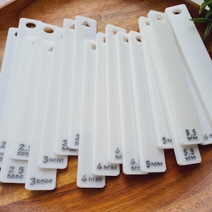 Peut inclure: Ensemble d'outils de jaugeage en acrylique blanc, chacun perc&eacute; et portant des mesures en millim&egrave;tres. Les outils sont dispos&eacute;s sur une surface en bois, avec des mesures allant de 2 mm &agrave; 6 mm.
