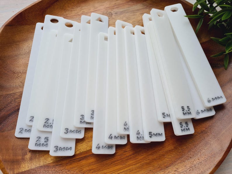 Peut inclure: Ensemble d'outils de jaugeage en acrylique blanc, chacun perc&eacute; et portant des mesures en millim&egrave;tres. Les outils sont dispos&eacute;s sur une surface en bois, avec des mesures allant de 2 mm &agrave; 6 mm.