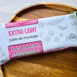 May include: A white package of self-hardening modelling clay labelled "EXTRA-LIGHT pâte de moulage" in pink lettering. The package is 160g and is labelled in four languages: French, Dutch, German, and English. The package also includes a description of the clay and its uses.