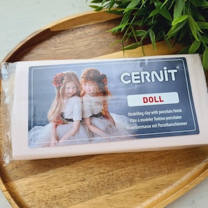 May include: A rectangular block of light pink Cernit Doll modelling clay with a porcelain finish, encased in clear plastic packaging. The label features a photograph of two children and the text Cernit Doll, Modelling clay with porcelain finish.