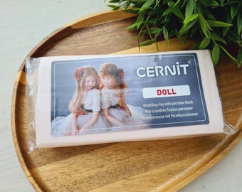 May include: A rectangular block of light pink Cernit Doll modelling clay with a porcelain finish, encased in clear plastic packaging. The label features a photograph of two children and the text Cernit Doll, Modelling clay with porcelain finish.