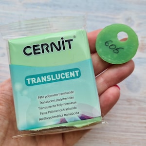 May include: A package of green translucent polymer clay by the brand Cernit. The package has the text "TRANSLUCENT" and the words "Pâte polymère translucide", "Translucent polymer clay", "Transluzente Polymermasse", "Pasta Polimerica traslucida", and "Arcilla polimérica translúcida" printed on it. A green circle with the number "605" written on it is next to the package.