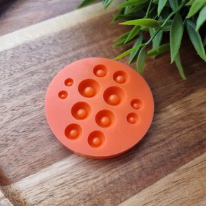 K&ouml;nnte beinhalten: Orangefarbene Silikonform mit 10 runden Vertiefungen in verschiedenen Gr&ouml;&szlig;en. Die Form befindet sich auf einer Holzoberfl&auml;che.